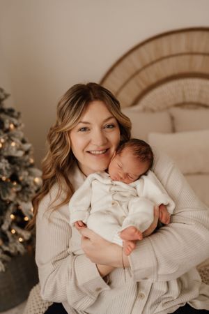 A parent in a cream sweater holds a sleeping newborn baby wearing white, with a Christmas tree in the background.