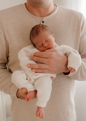 A sleeping newborn baby wearing white knit clothing is cradled gently in someone's arms.