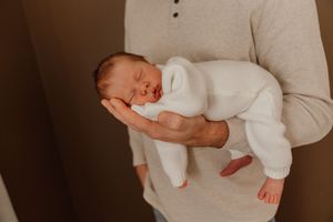 A sleeping newborn baby wearing white clothing is cradled in gentle arms against a neutral background.