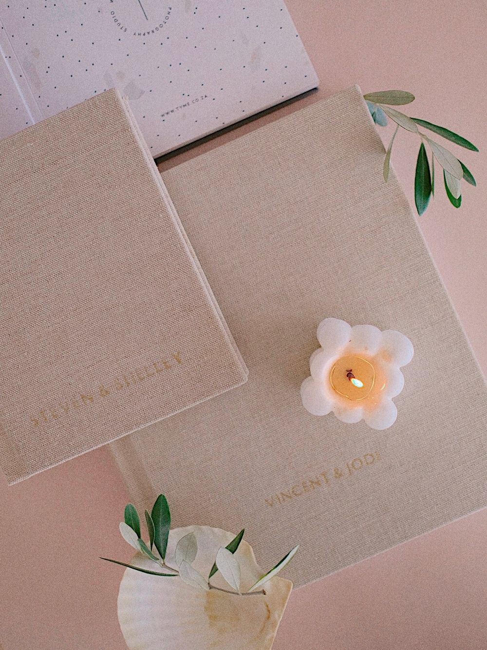 Two linen-covered wedding guest books with gold foil names, adorned with a flower candle and olive branches on pink background.