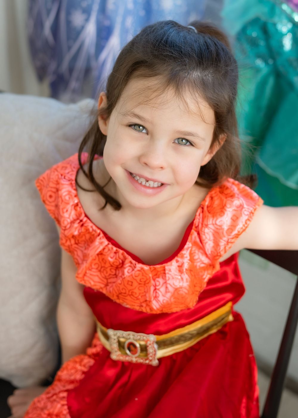 Young child wearing a bright red and coral dress with ruffled neckline and gold belt sitting on beige couch.