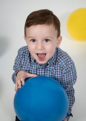 Child in blue checkered shirt poses playfully with a bright blue balloon against a white background.