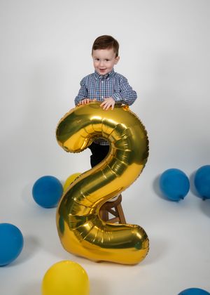 Child plays with a large gold number 2 balloon during a birthday photo session with blue and yellow accents.
