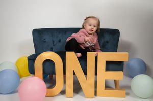 Baby sits on blue couch with colorful balloons and gold ONE letters for first birthday photoshoot celebration.