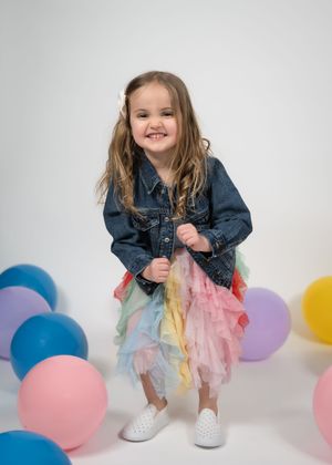 Young child in denim jacket and colorful tulle skirt poses playfully among pink, blue and yellow balloons on white background.