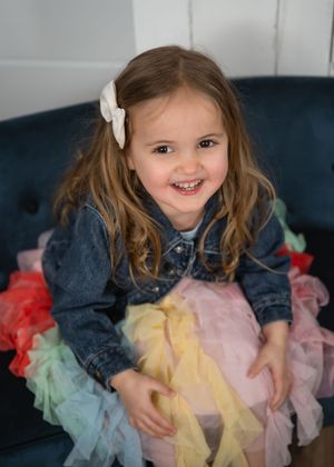 A young child wearing a denim jacket and colorful tulle skirt smiles brightly while sitting on a dark blue couch.