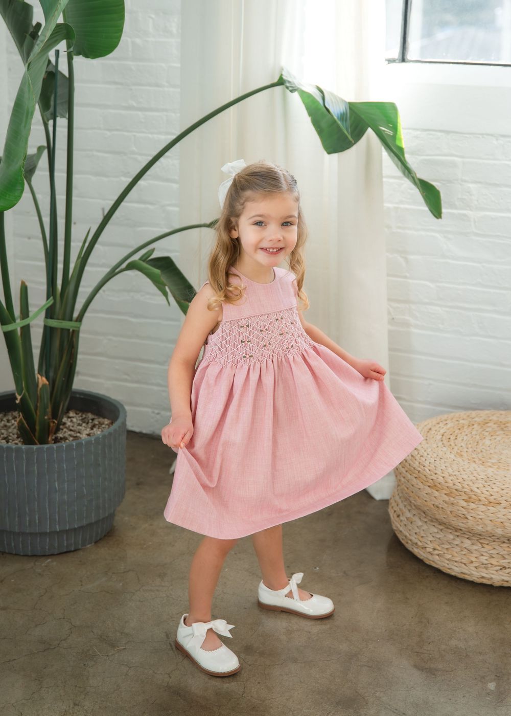 A young child twirls in a pink summer dress and white shoes next to a large potted plant in a bright indoor setting.