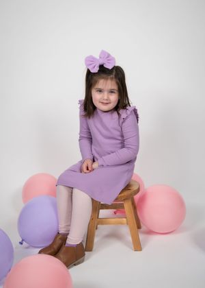 A child in a purple dress sits on a wooden stool surrounded by pink and purple balloons against a white backdrop.