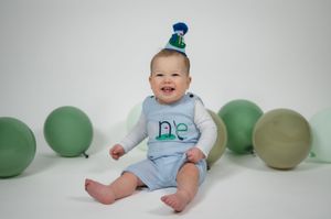 Baby in light blue outfit sits and smiles while surrounded by sage green balloons in a minimalist studio setting.