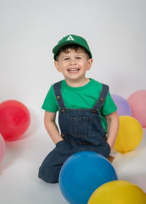 A cheerful figure in overalls and a green baseball cap poses playfully against a white background with colorful balloons.
