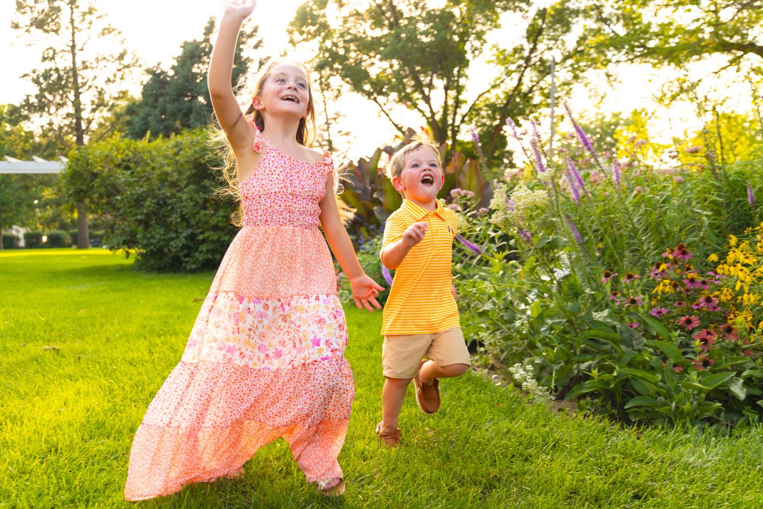 Two children dance and play joyfully together in a sunny garden with flowers during golden hour sunset.
