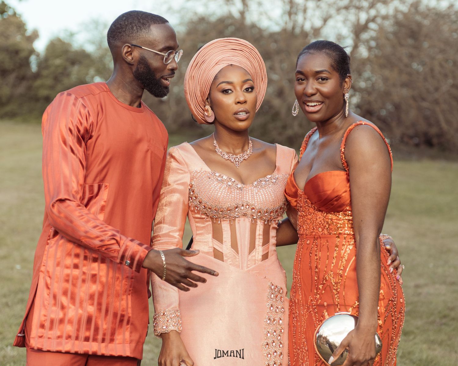 Bride in peach Nigerian traditional lace dress poses with man and woman in orange outfits outdoors.
