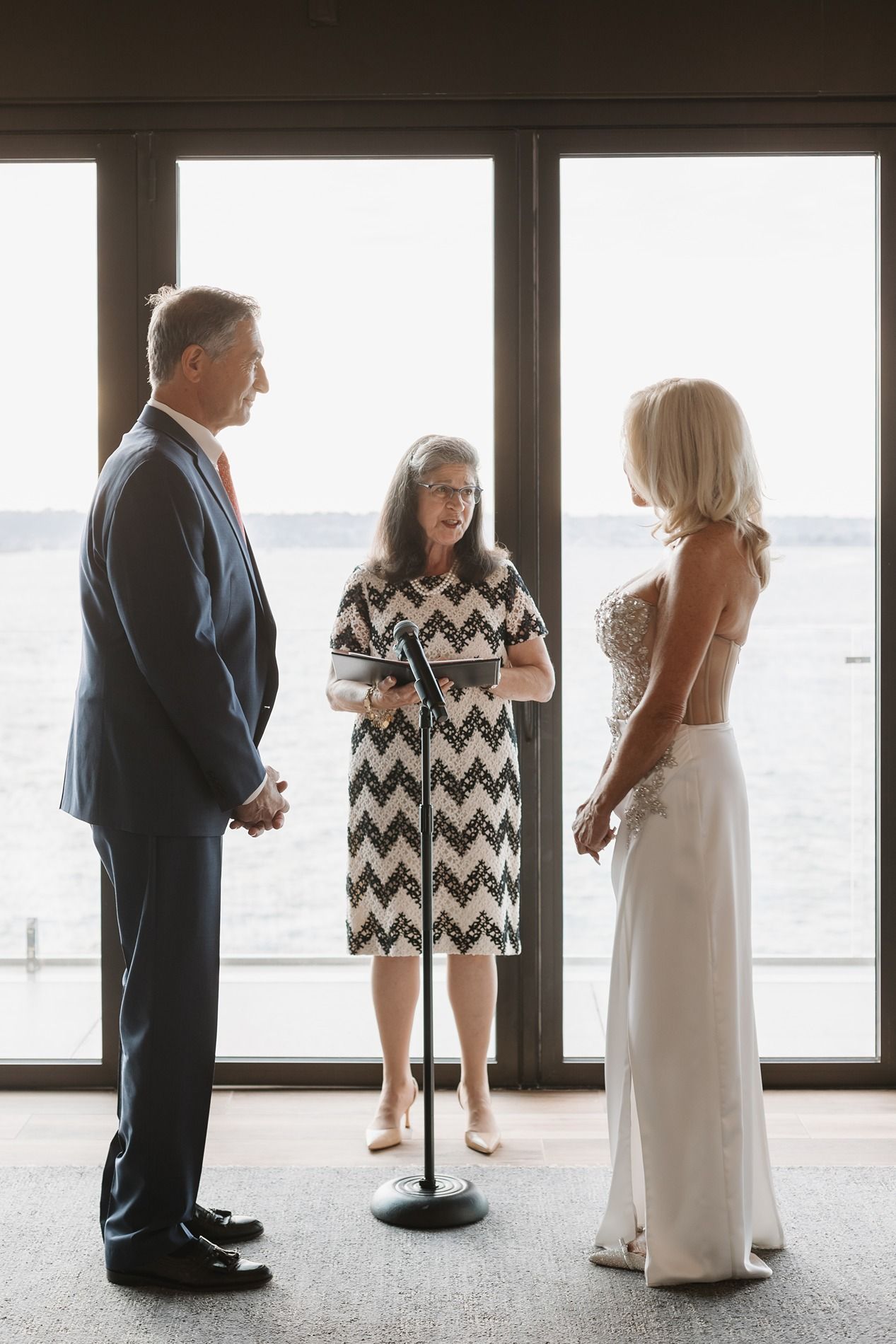A bride and groom stand in silhouette against large windows overlooking a waterfront view at an elegant venue.