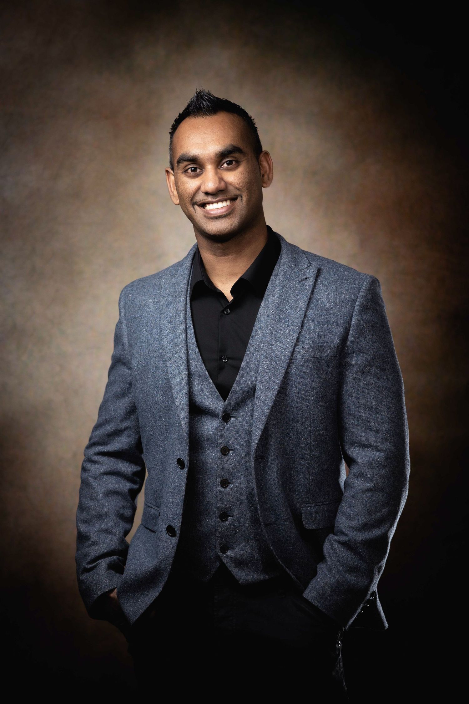 Professional studio portrait of a person in a gray three-piece suit with a black shirt against a brown background.