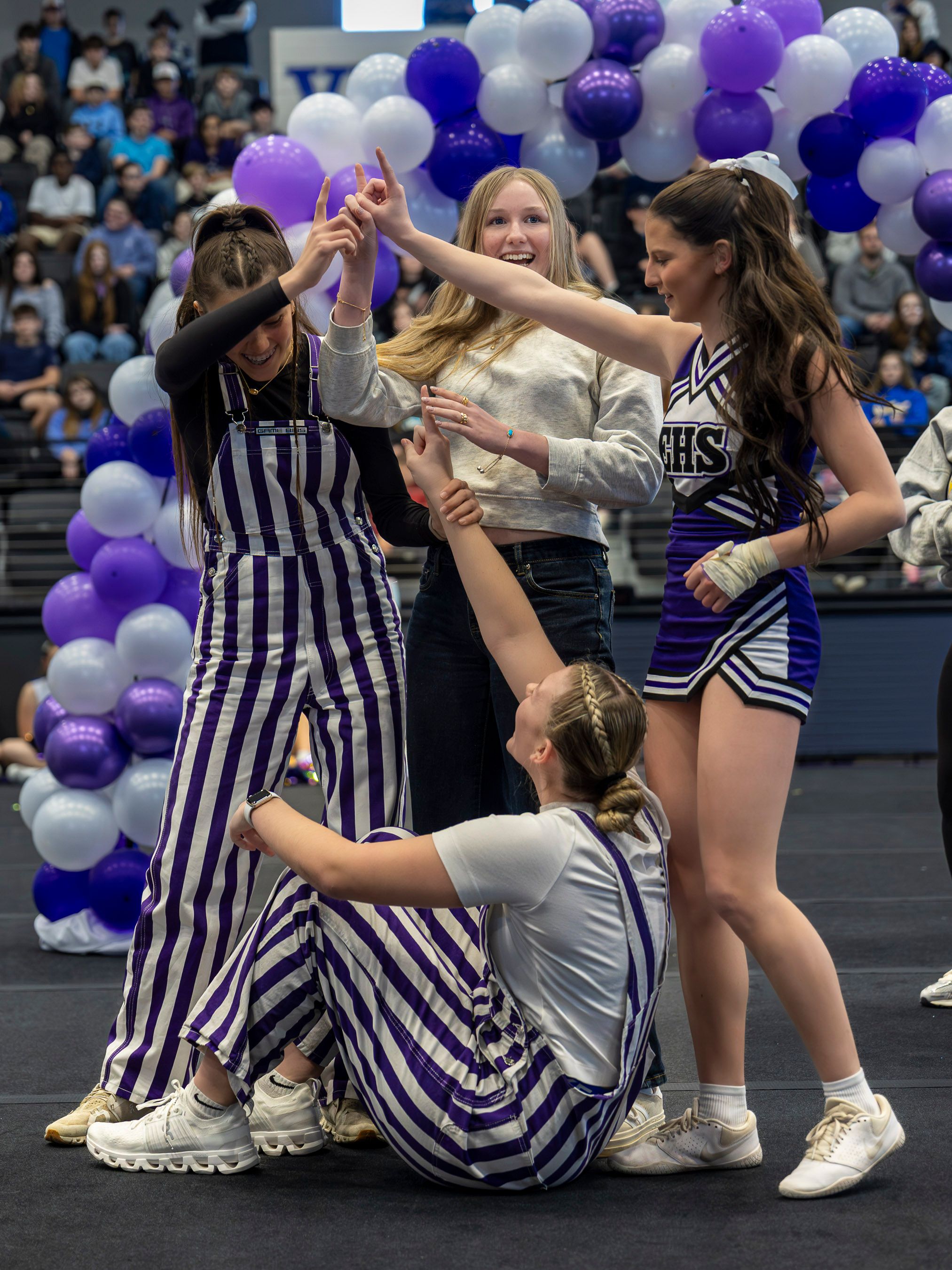 Cheerleaders in striped uniforms perform with purple and white balloons decorating the indoor sports venue.