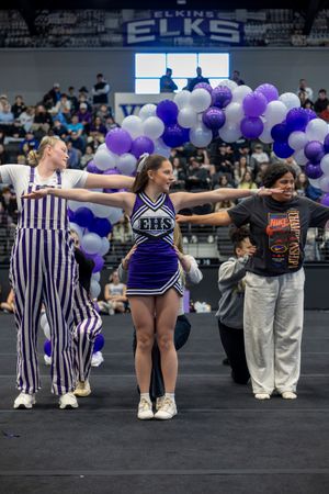Performers in striped outfits and cheer uniforms pose during a routine with purple and white balloon decorations.