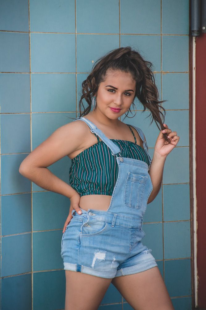 A person poses playfully in denim overalls and striped crop top against a turquoise tiled wall.