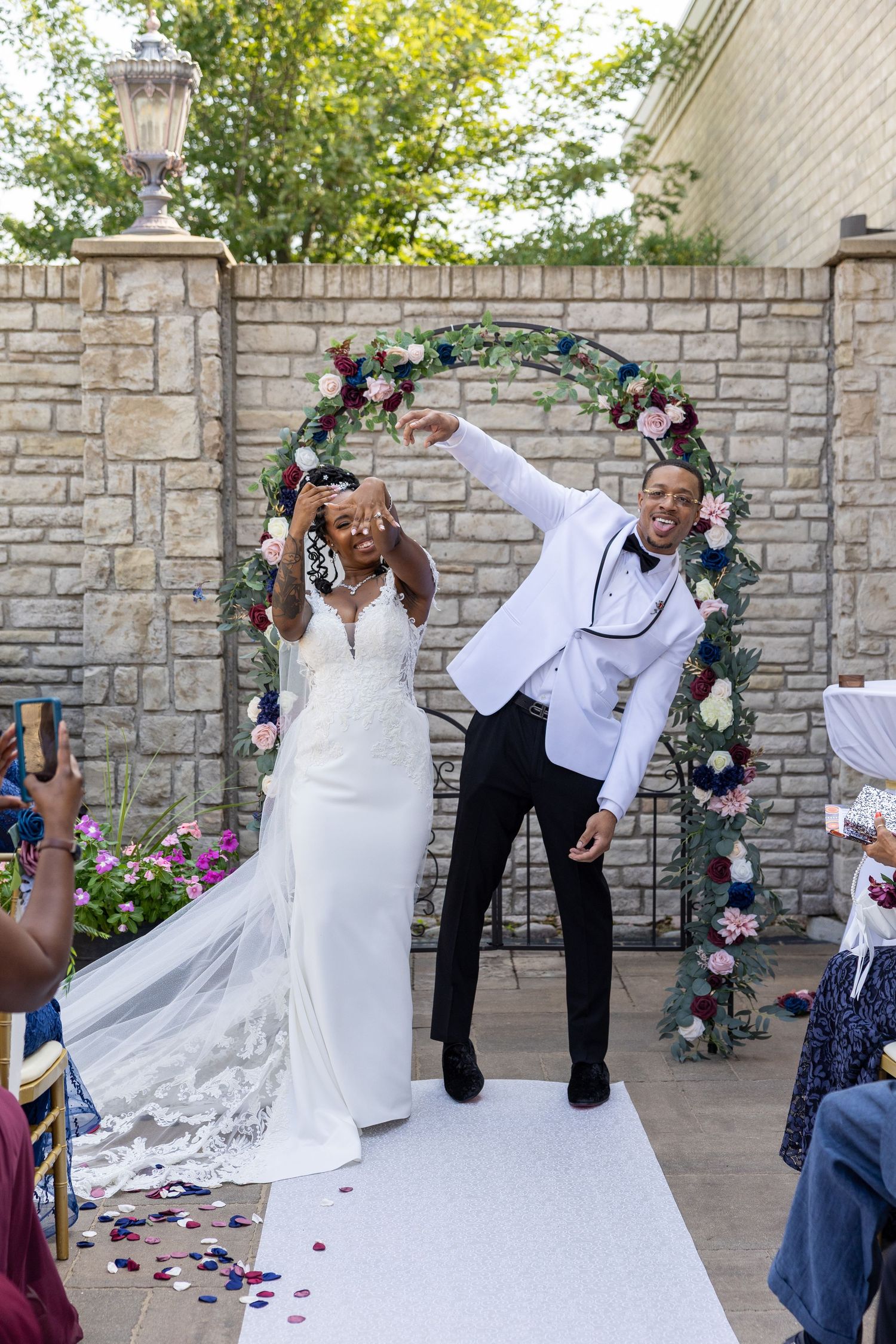 A wedding couple performs a fun dip pose during their outdoor photo session by a stone wall with floral decorations.