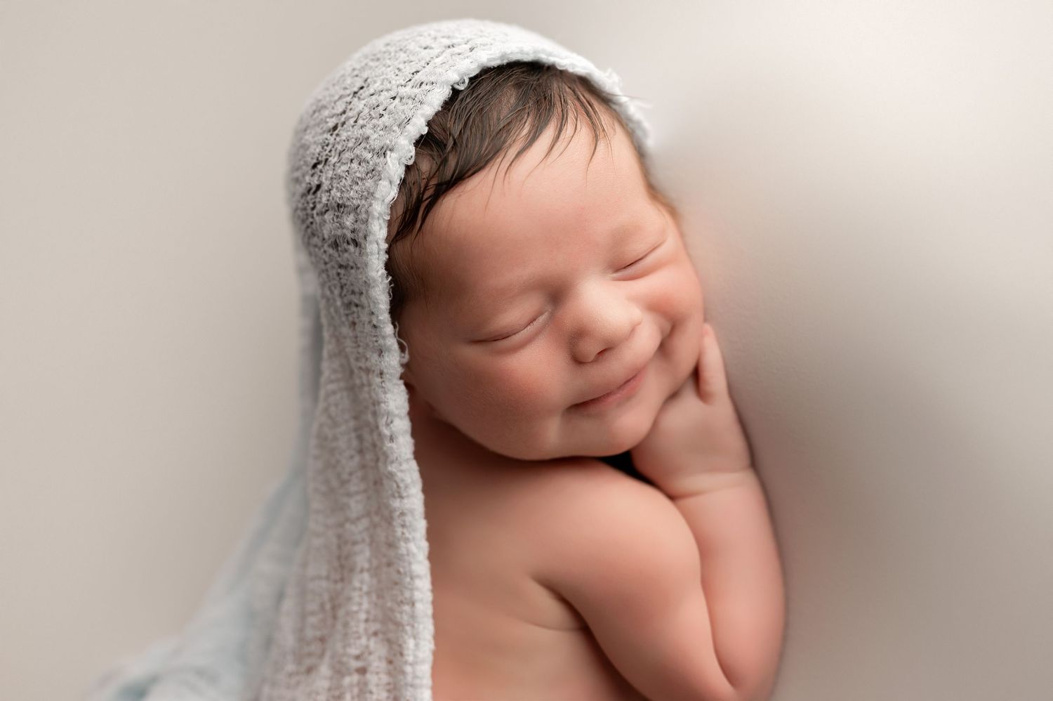 Peaceful sleeping newborn baby with soft smile wearing knitted white bonnet against neutral background.