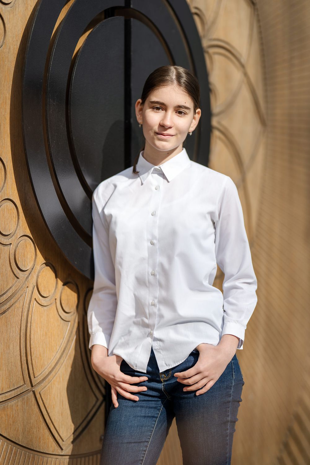 A person in a white button-down shirt and jeans poses against a tan wall with decorative circular designs.