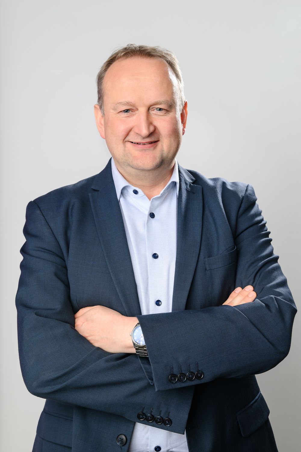 Professional business portrait of person in navy suit with white shirt against gray background.
