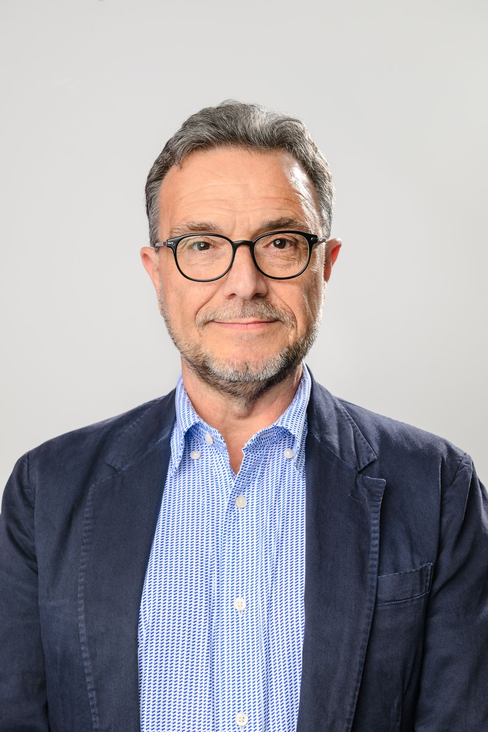 Professional headshot of person wearing glasses and navy blazer against gray background.
