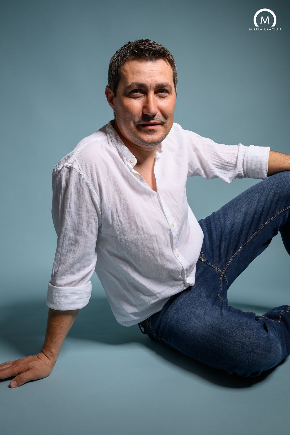Studio portrait of a man in white shirt and jeans against blue backdrop.
