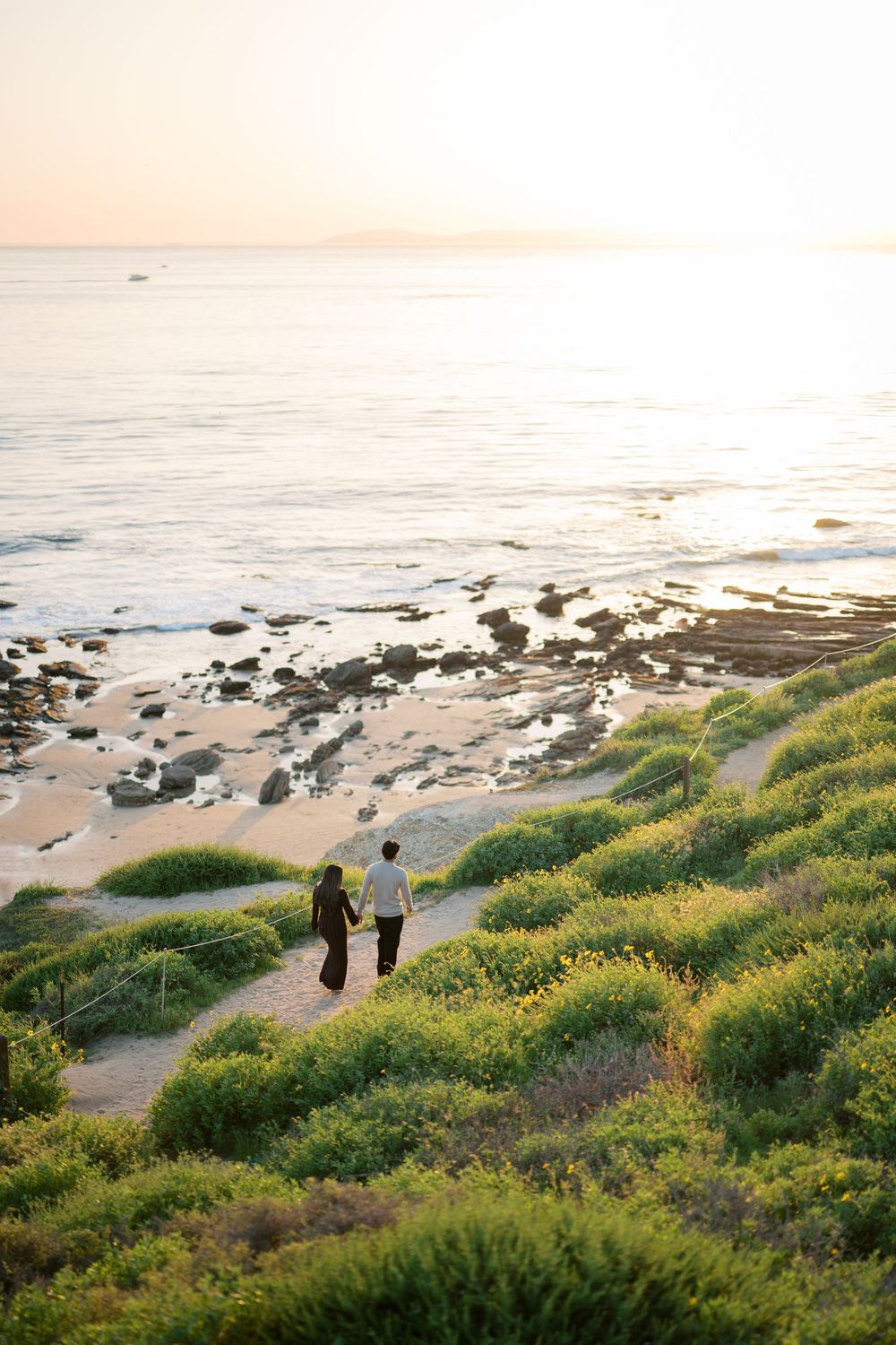 Two figures walk along a coastal path with wildflowers and ocean views at sunset.