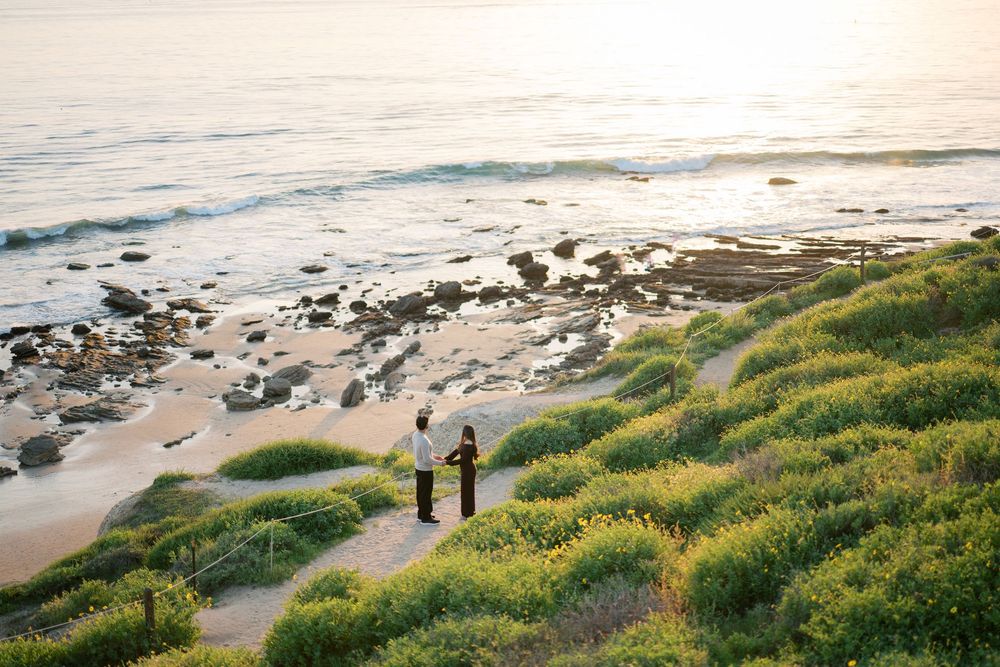 Couple walking along scenic coastal path with green grass and rocky beach during golden sunset hour.
