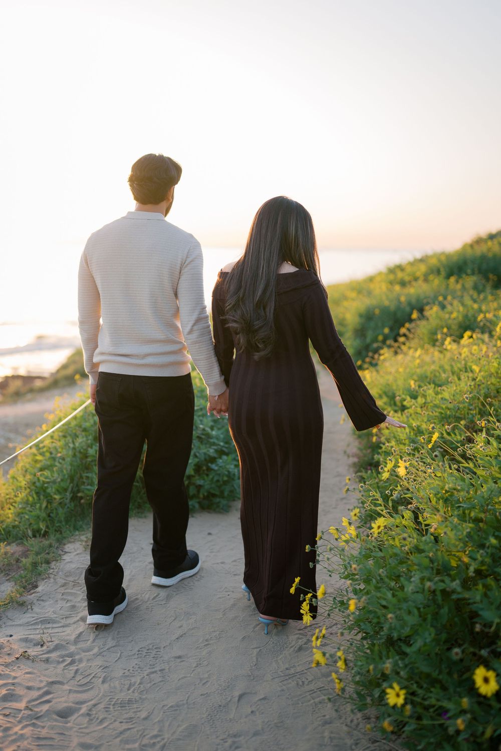 A couple walking hand in hand on a coastal path at sunset surrounded by yellow wildflowers.