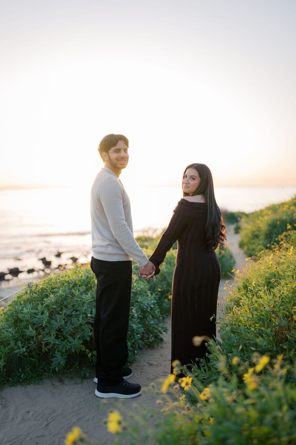 A romantic couple holds hands while standing on a seaside trail with yellow flowers at golden hour.