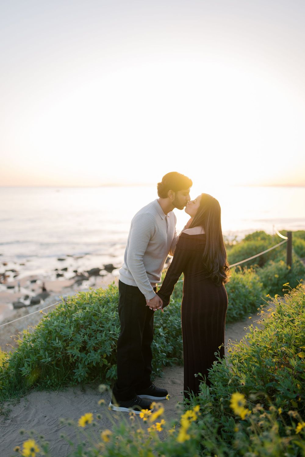 A couple shares an intimate moment among yellow wildflowers on a coastal cliff at sunset.