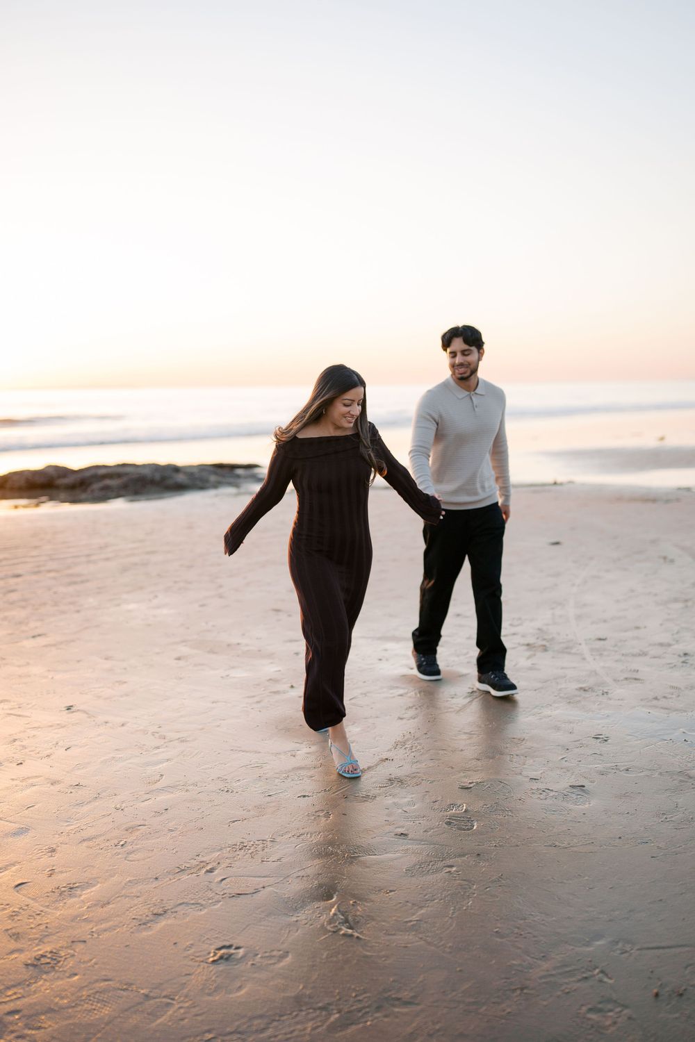 Two people walk together on a sandy beach at sunset, leaving footprints in the wet sand.