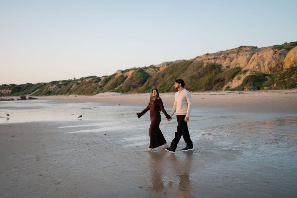 A couple walks hand in hand along a scenic beach with dramatic cliffs in the background during golden hour.