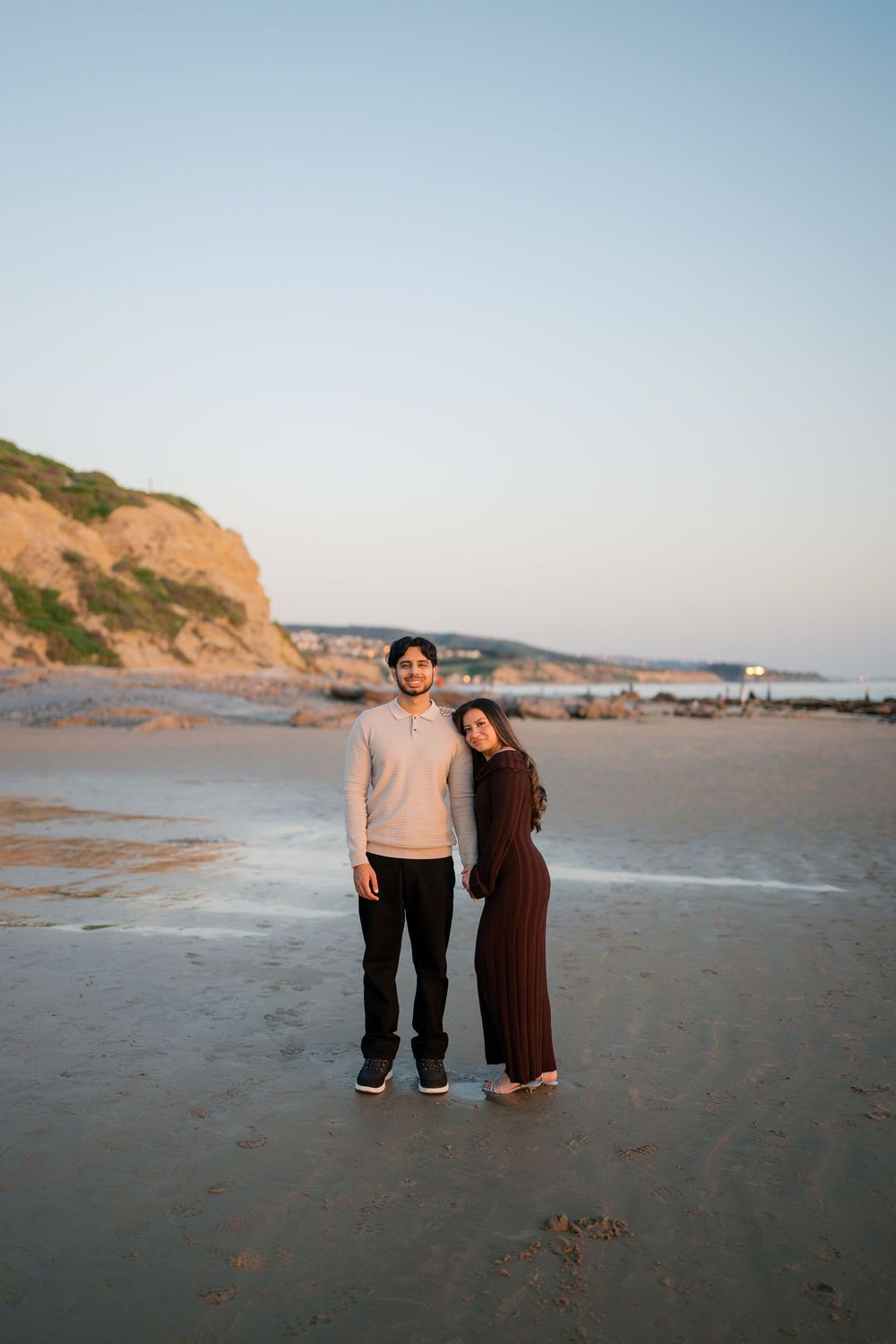 A couple stands together on a sandy beach with coastal cliffs in the background during golden hour.