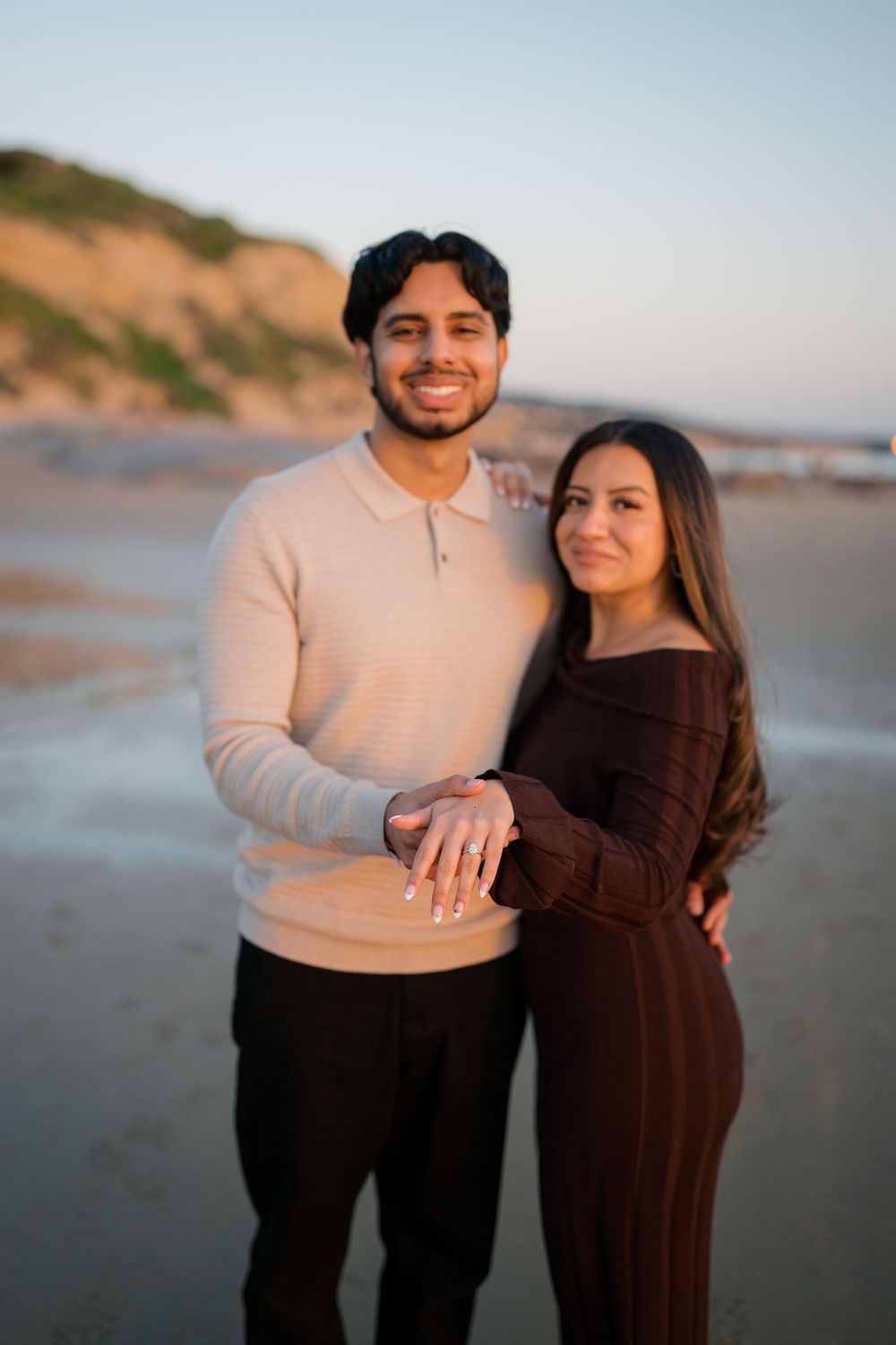 A couple embraces on a misty beach shoreline with cliffs and ocean waves visible in the distance.