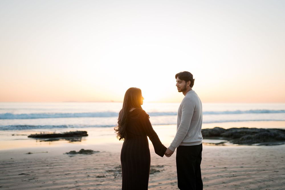Two silhouettes holding hands while watching sunset on a peaceful beach with waves rolling in.
