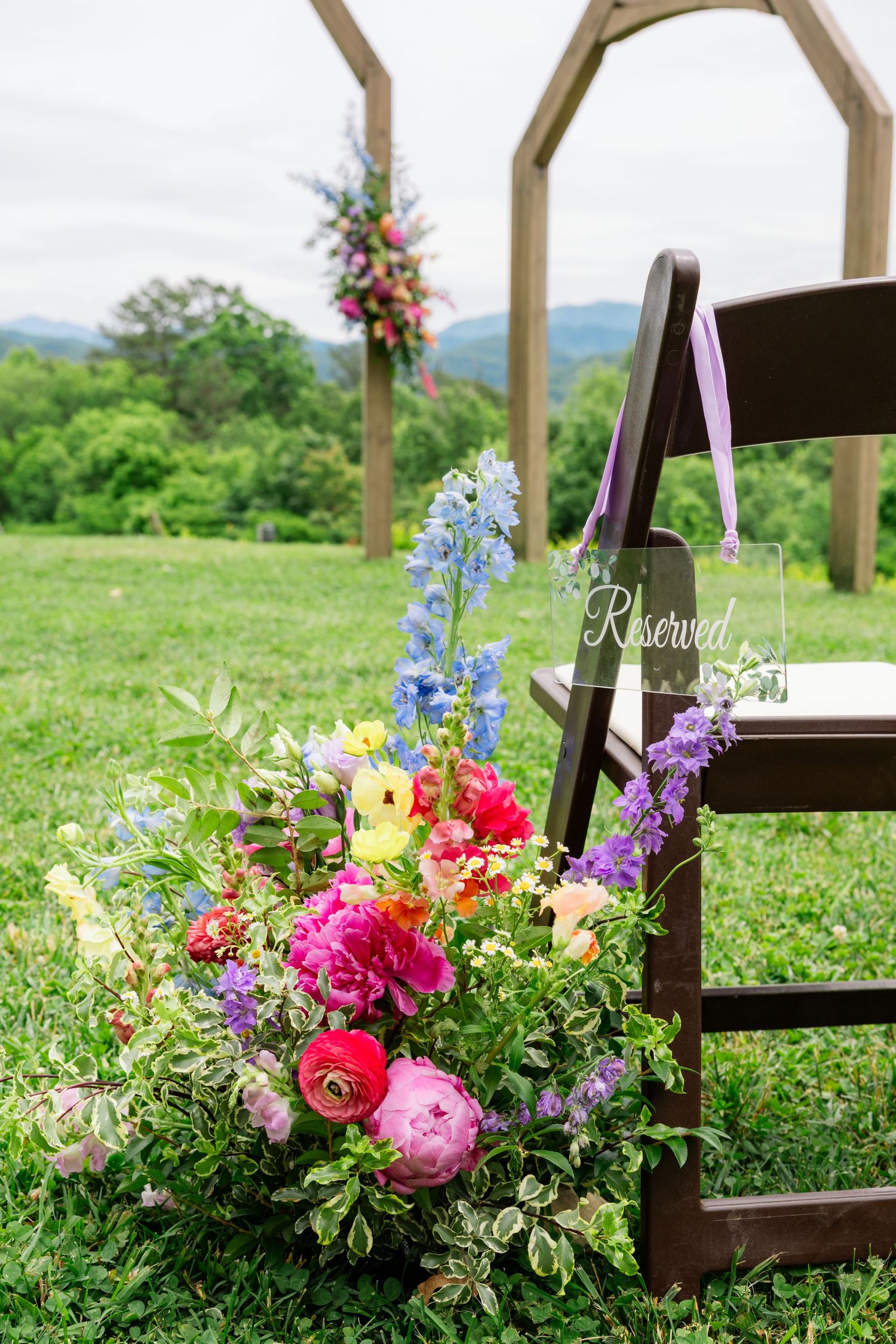 Colorful floral arrangements with pink, purple, and yellow blooms decorate wooden chairs at an outdoor wedding venue.