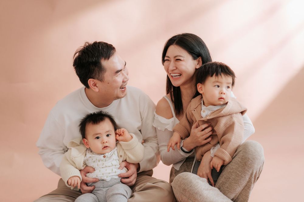 Family in matching beige outfits shares joyful moments during an indoor photoshoot with soft pink lighting.