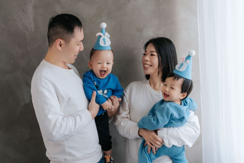 Family wearing matching white and blue outfits laugh together against gray wall during indoor portrait session.
