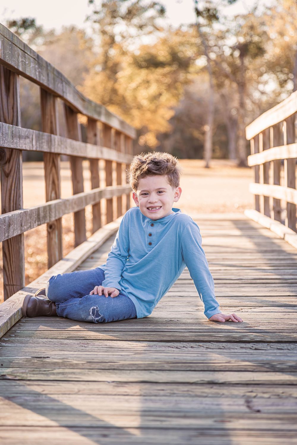 Child lying on wooden boardwalk bridge during golden hour sunset creating playful outdoor portrait series.