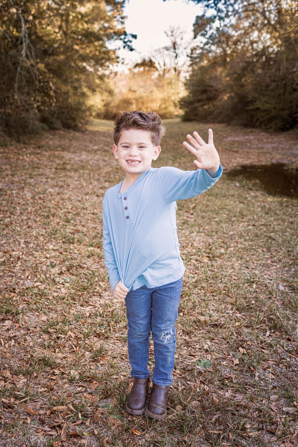 A young person in a blue henley shirt and jeans waves at the camera while standing on a leaf-covered path in autumn.