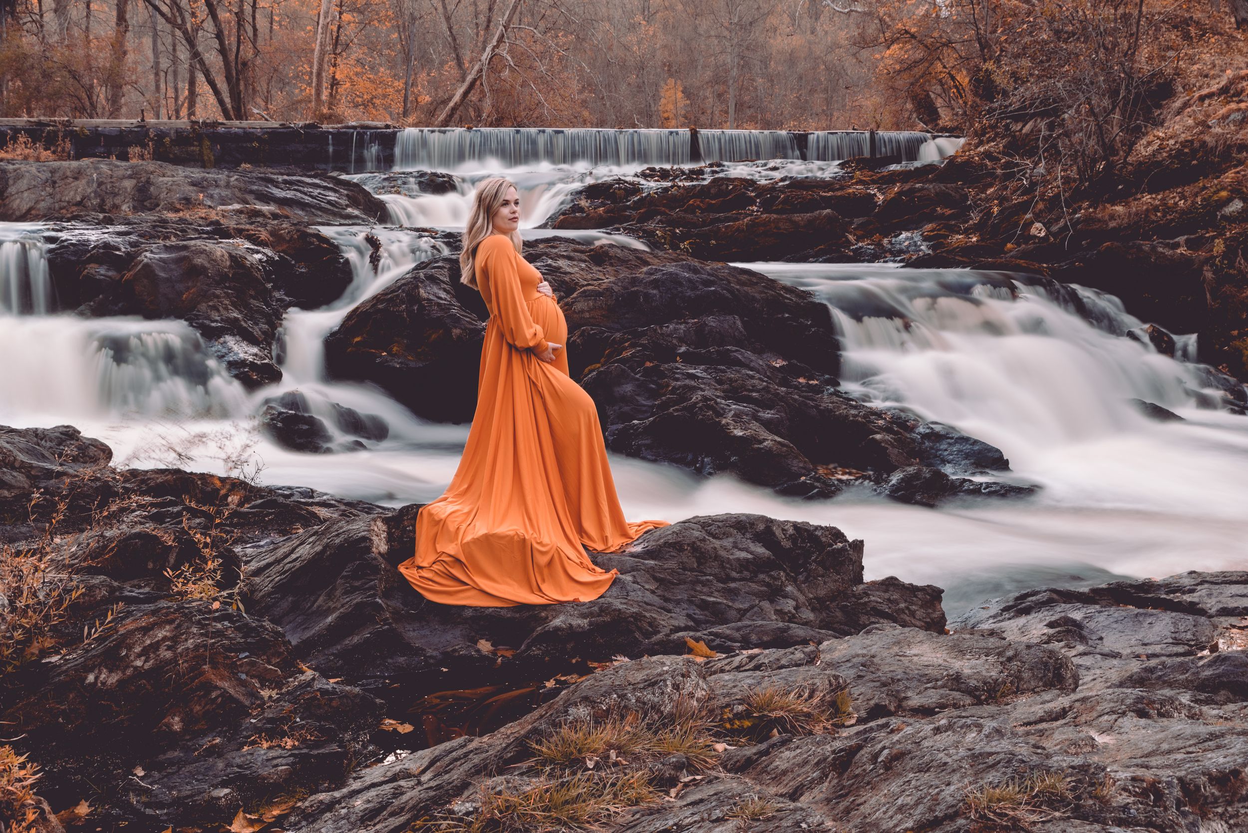 Person in orange dress sitting on rocks by cascading waterfall in autumn setting.