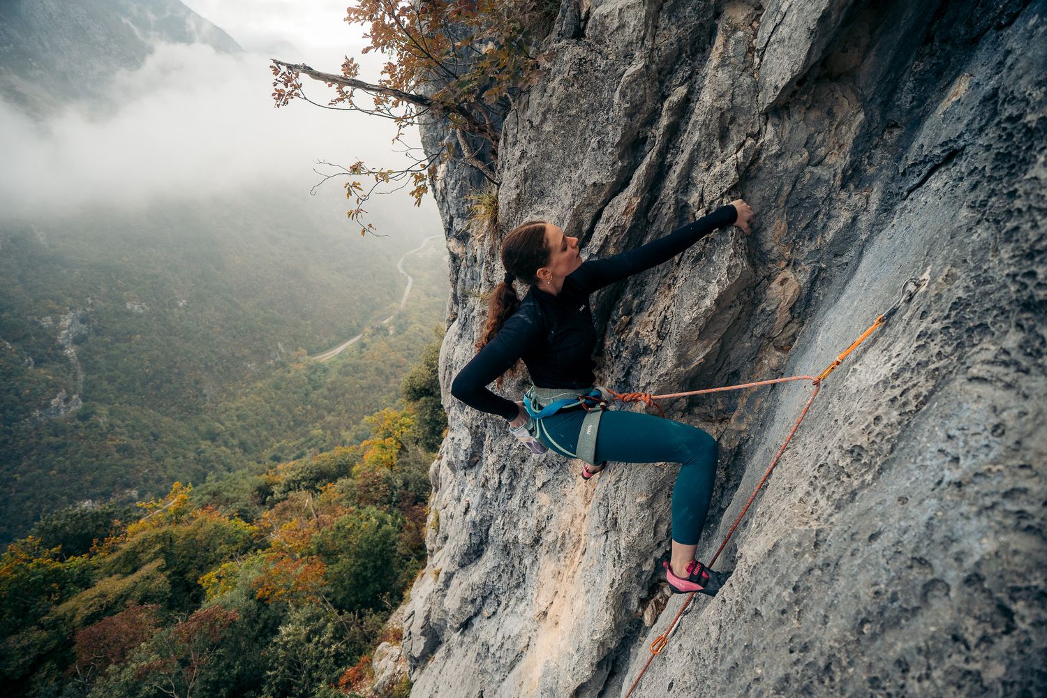 Rock climber ascending a steep limestone cliff face with autumn foliage visible in the misty mountain background.
