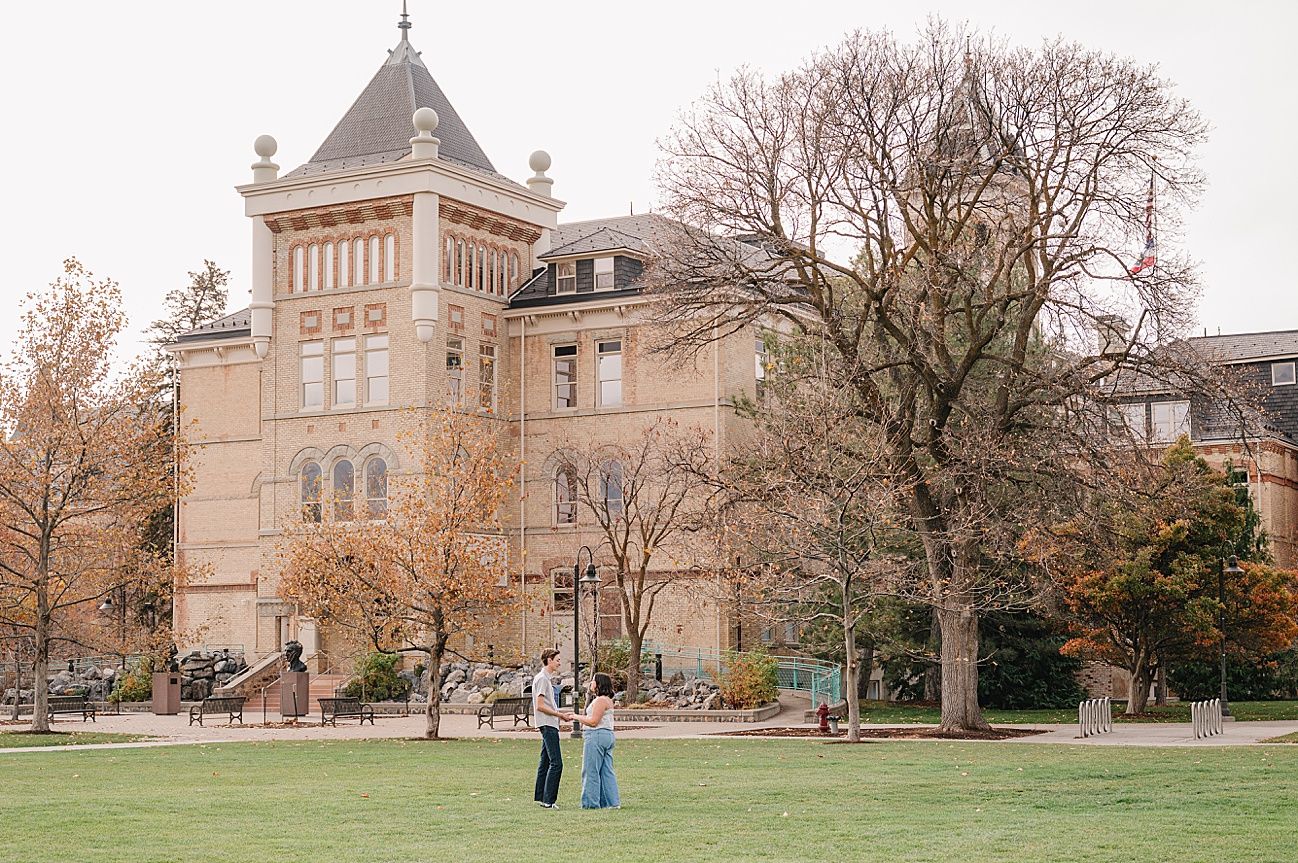 A couple in formal attire stands on a grand lawn in front of a historic mansion during an autumn wedding photo session.