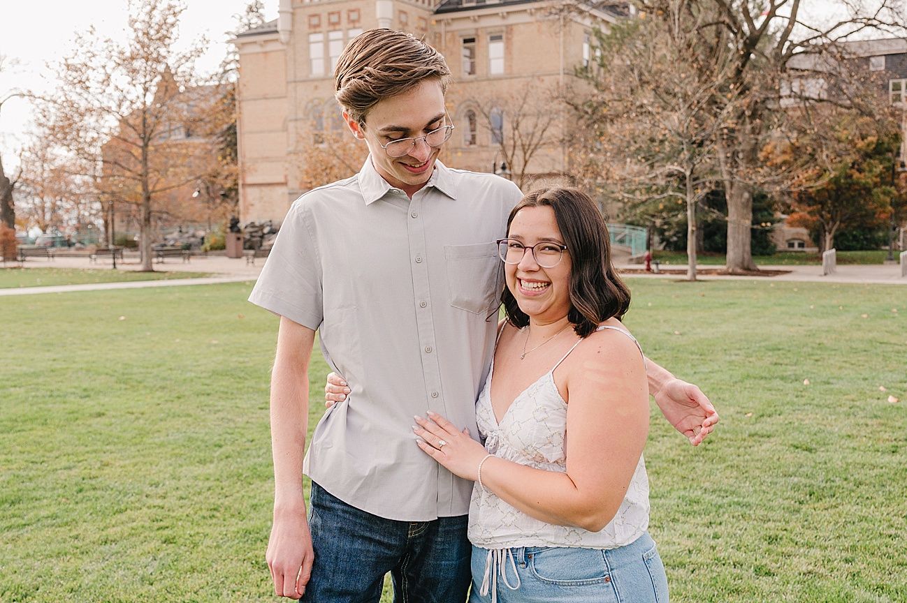 A sweet proposal moment unfolds on a college campus lawn with historic architecture in the background.