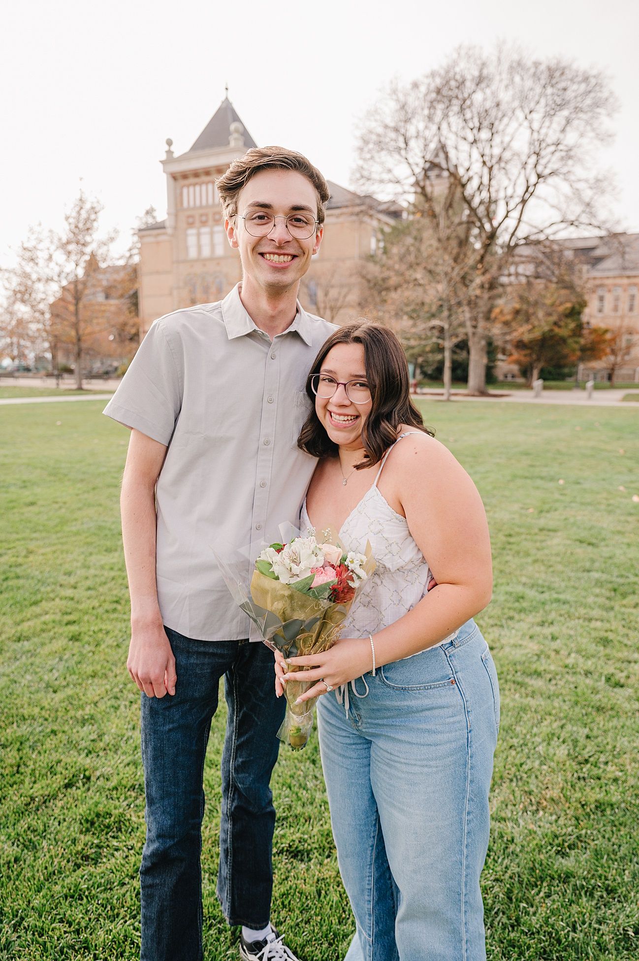 A couple celebrates their engagement while holding flowers on a grassy campus lawn.