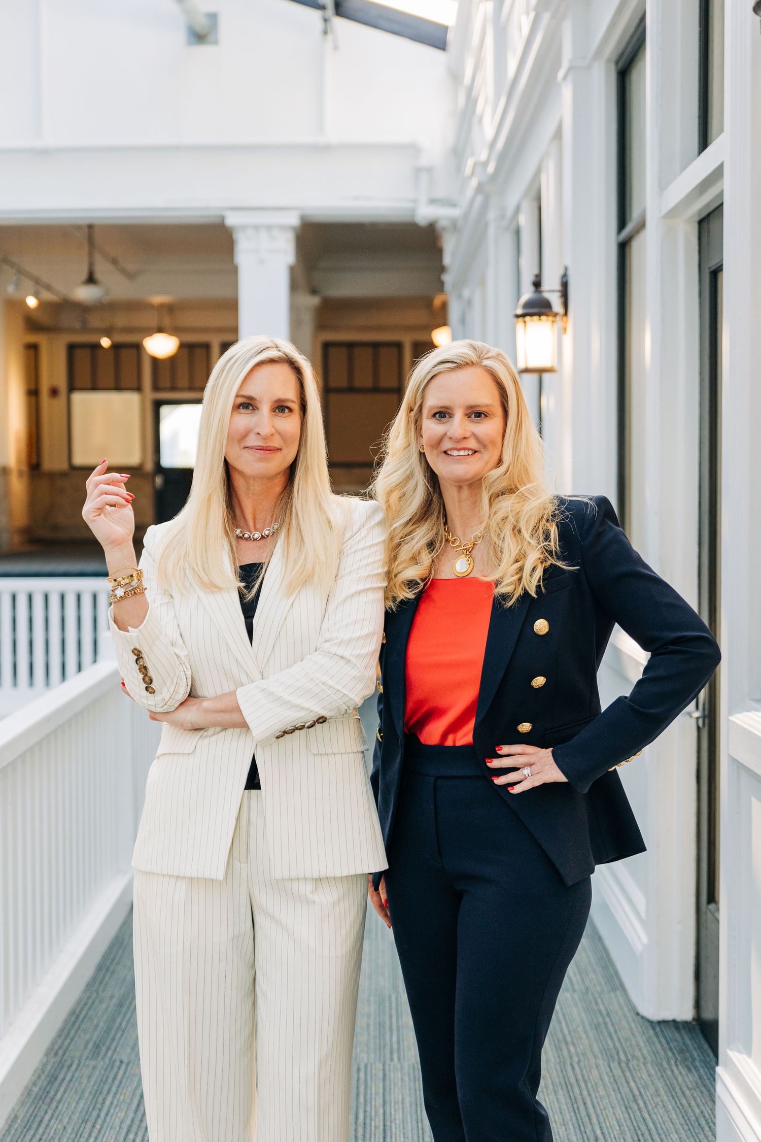 Two professional businesswomen in contrasting suits stand confidently in a bright hallway with white columns and lighting in Charlotte