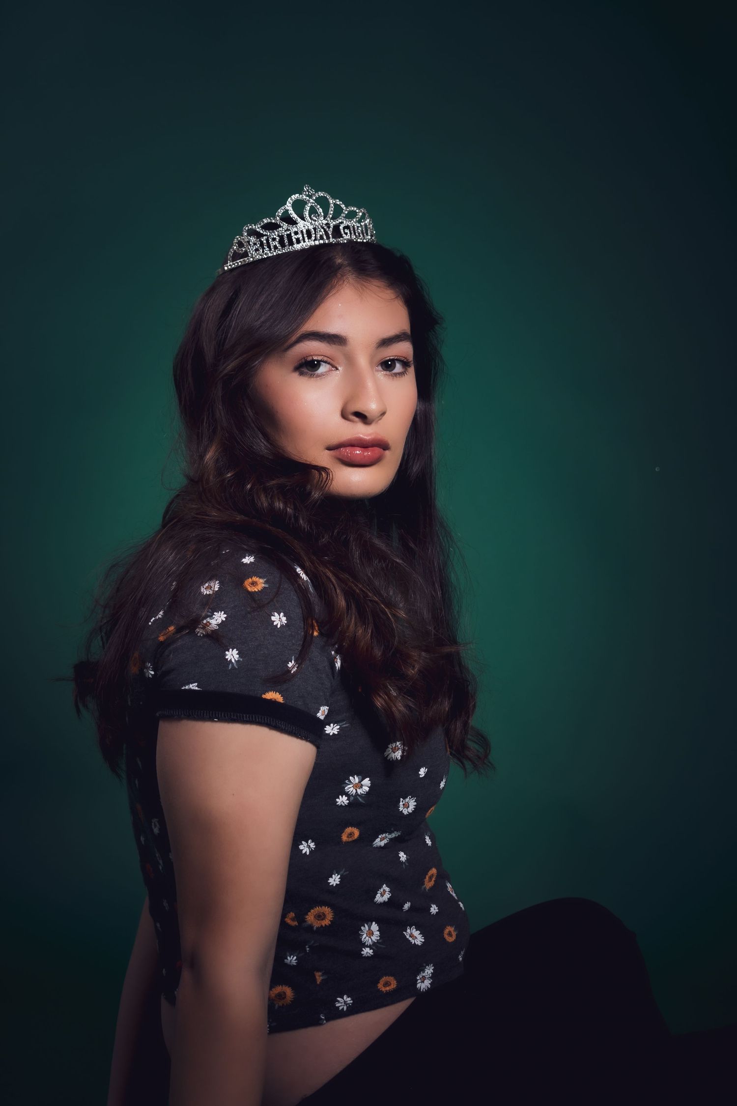 Studio portrait with dark green background showing someone wearing a black floral dress and small tiara.