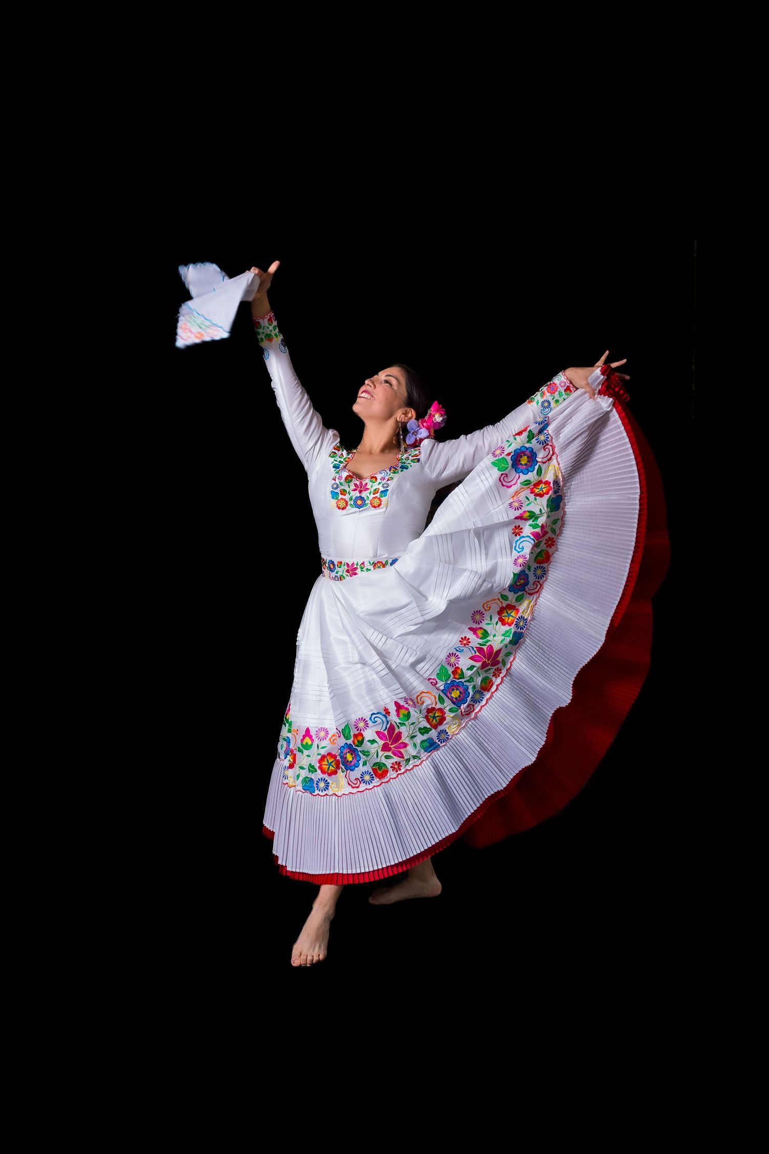A dancer in a traditional white Mexican dress with colorful floral embroidery performs graceful spinning movements against black.